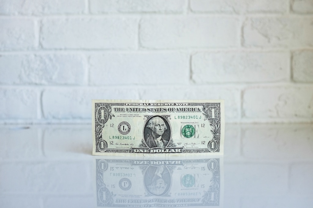 A US one dollar bill standing upright on a white shiny desk against a white brick wall