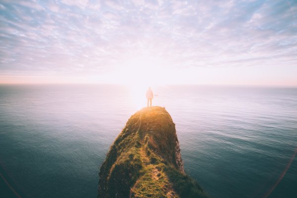 A person standing on a cliff jutting out into the ocean with the sunrise in front of them