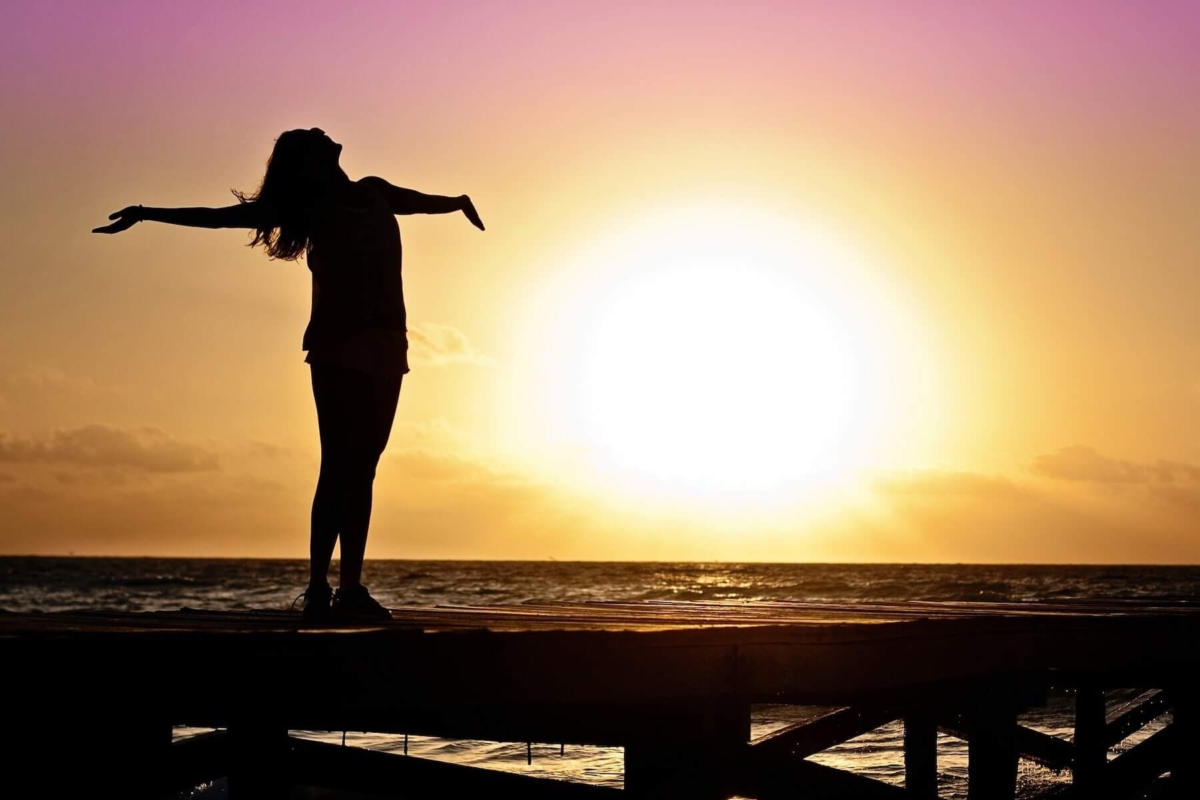 A woman standing on a pier jutting out into the water with her head back and her arms thrown wide. An orange and pink sunset in the background