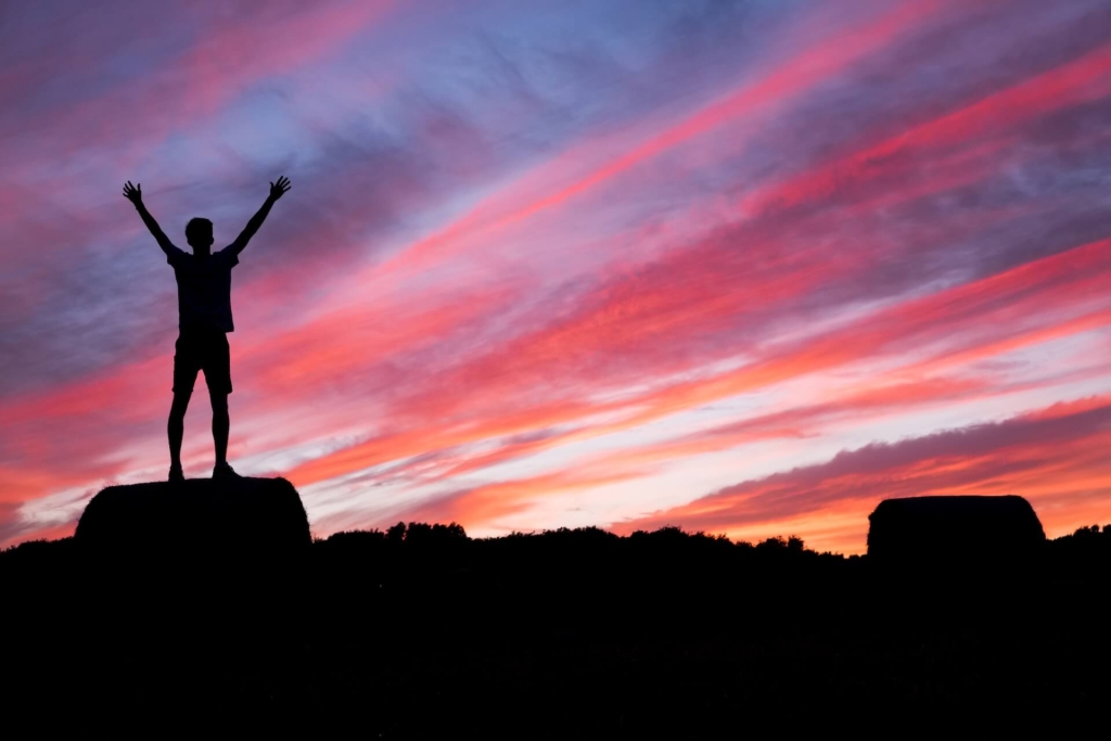 The silhouette of a man with his arms in the air standing on a hill in front of a colorful sunset